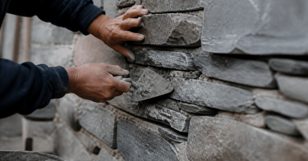 Mains appliquant mortier entre pierres grises alignées horizontalement
